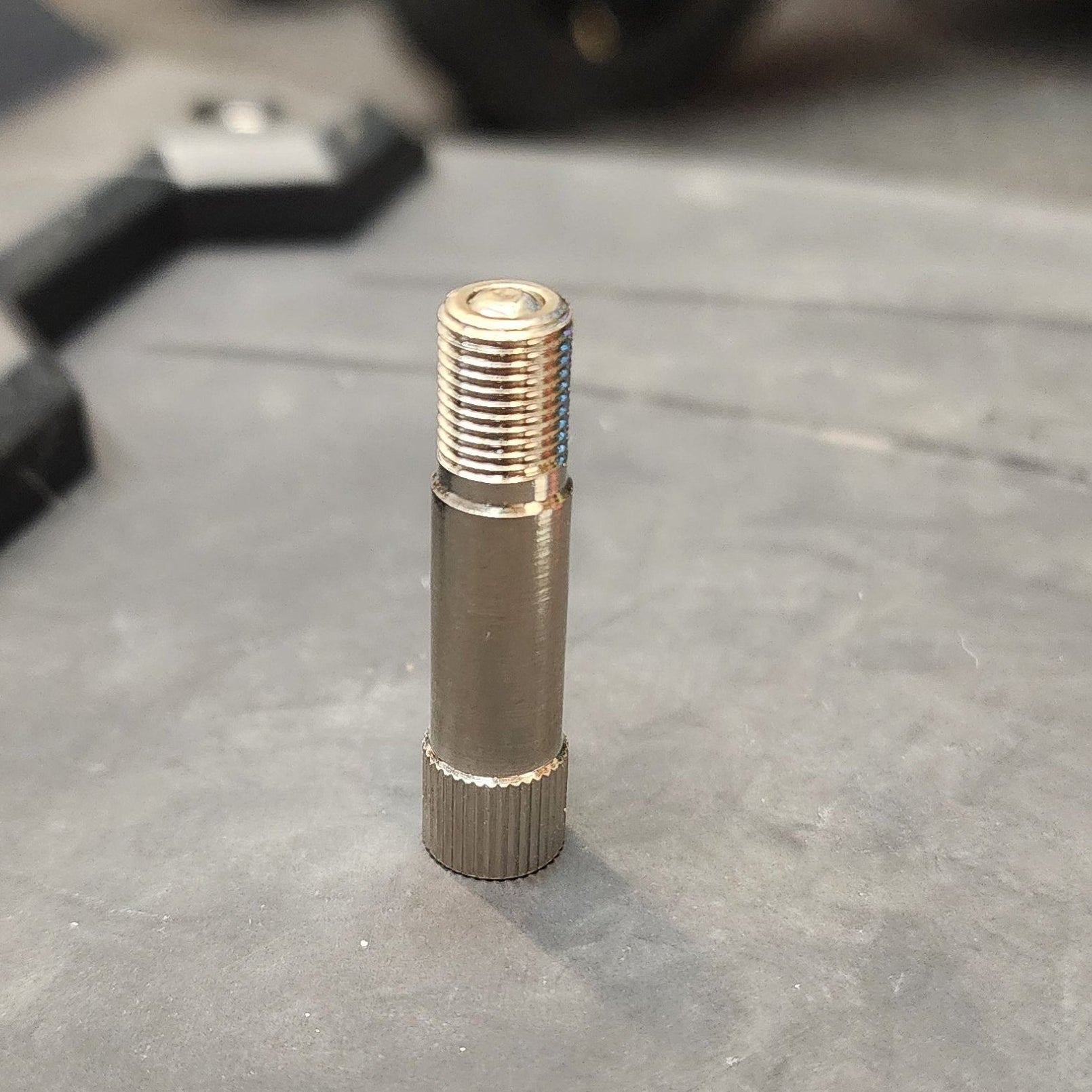 A vertical, cylindrical chrome tire valve extender with a threaded top and a knurled base, standing on a textured grey surface with blurred vehicle wheels in the background.