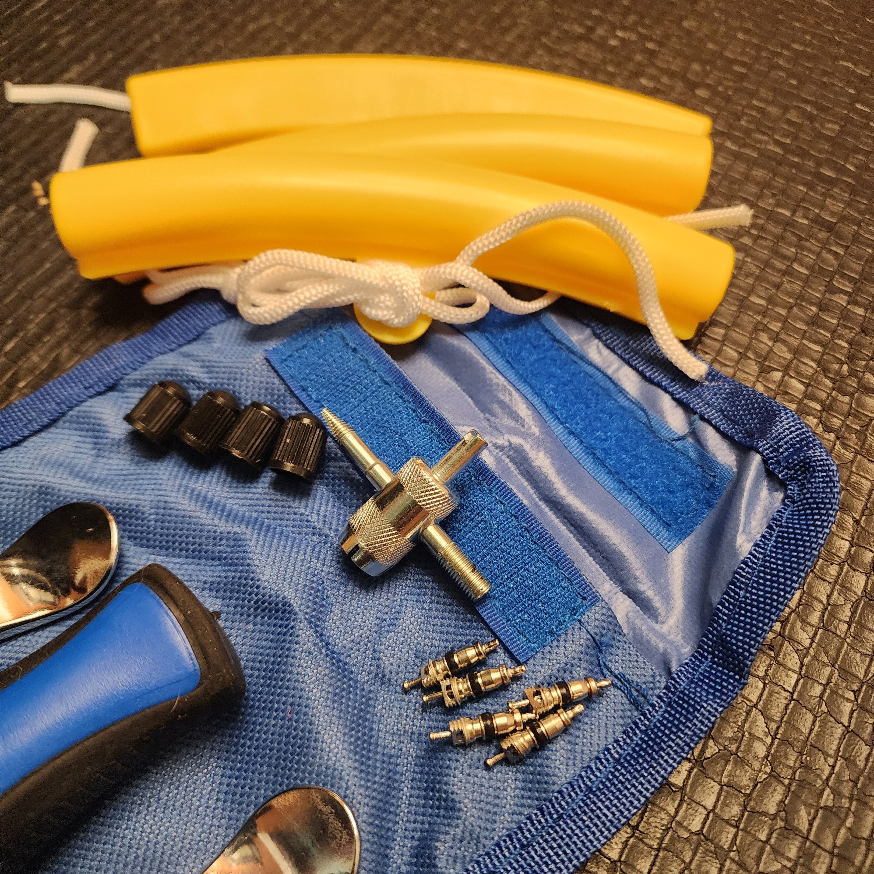 A close-up of bicycle inner tube repair tools laid out on a textured blue canvas bag, yellow rim protectors, a blue handled tire lever, metal valve tools, and several valve stem caps